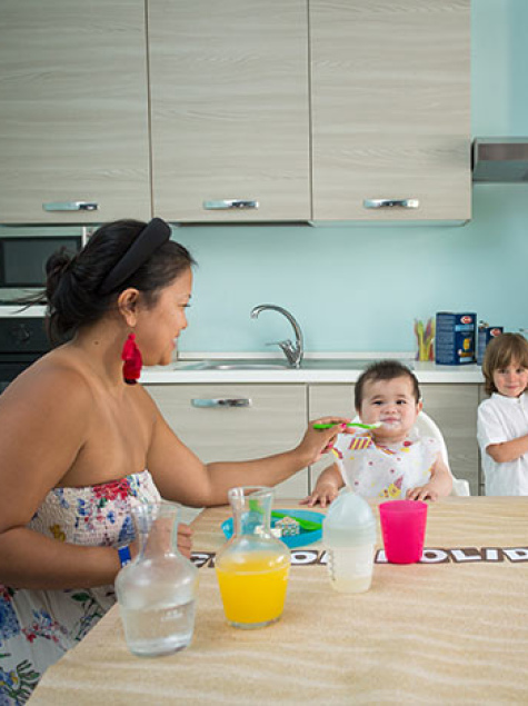 Famiglia in cucina, madre prepara, bambino sorride, altra donna dà da mangiare al piccolo.